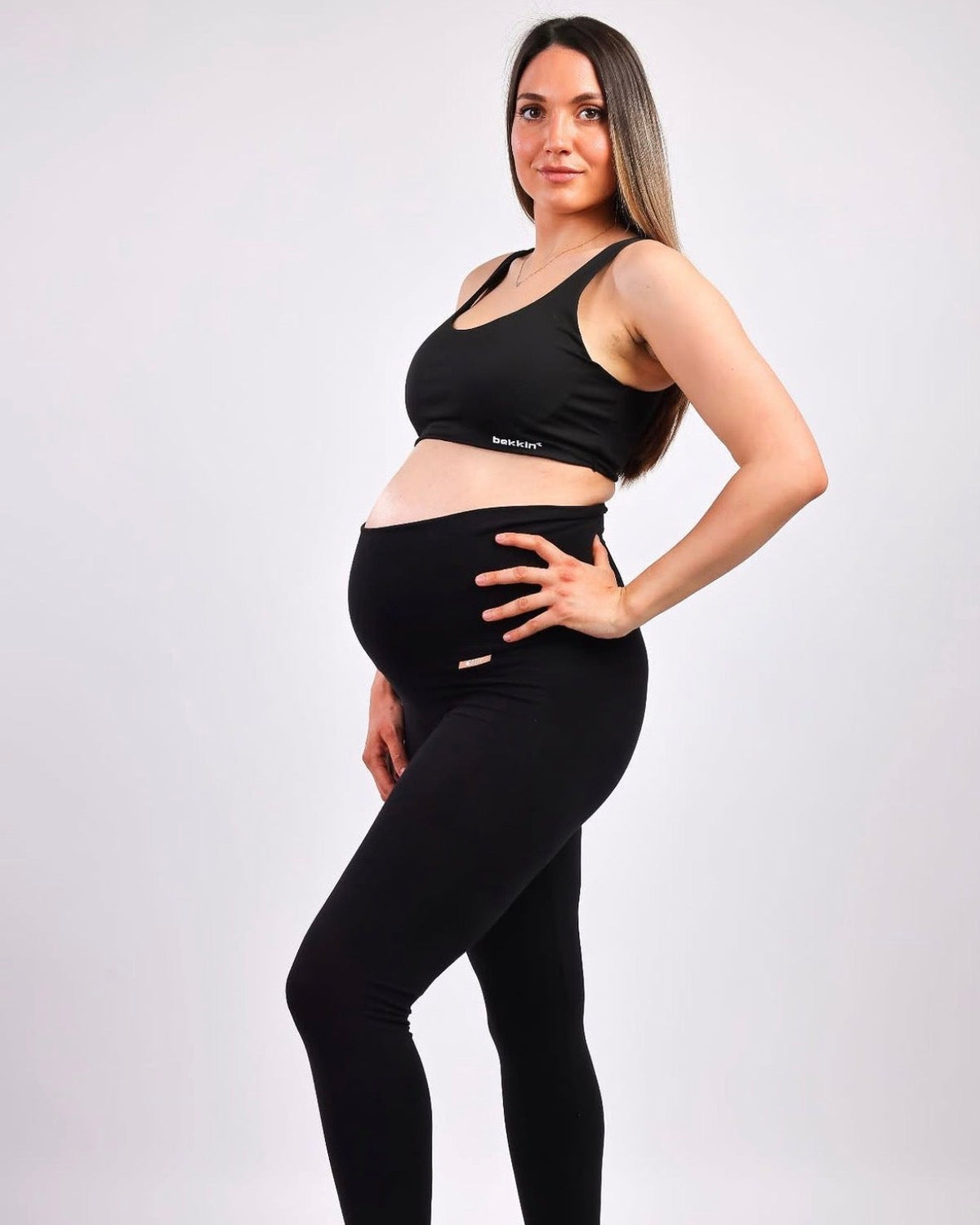 A pregnant woman posing in a black sports bra and matching high-waisted maternity leggings against a light gray studio background.